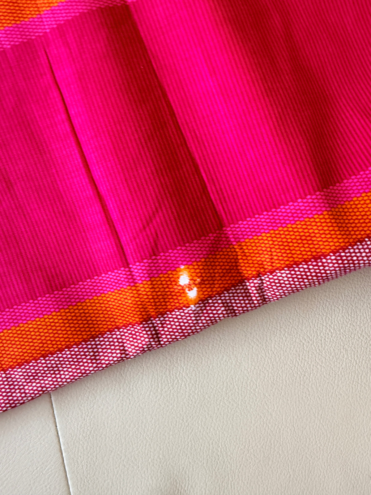 A close-up of a bright pink fabric with vertical texture and a striped border in pink, orange, and white, laid on a light beige surface.