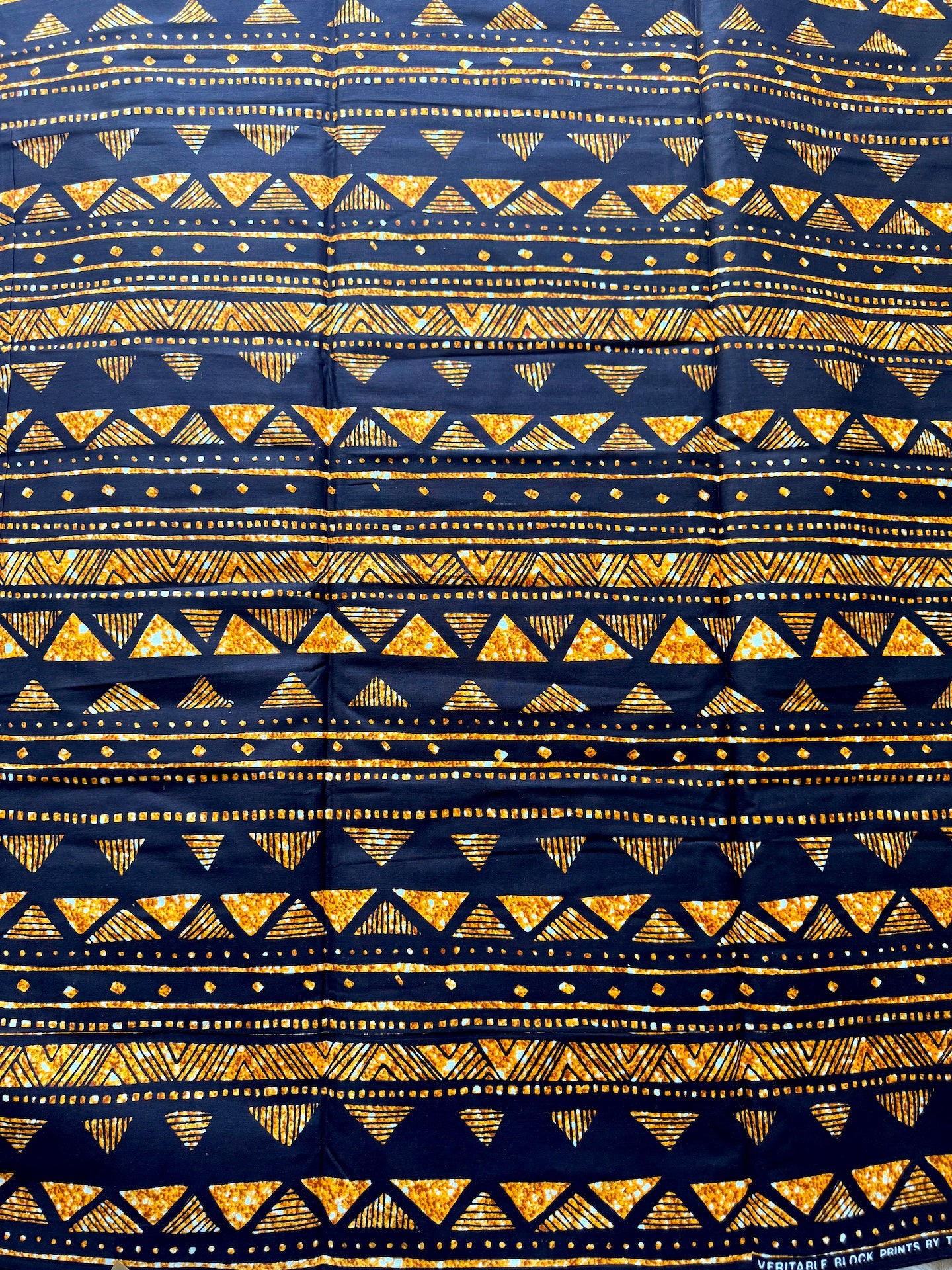 A close-up of a navy blue fabric with a repeating geometric pattern of yellow and gold triangles, dotted lines, and hatched stripes.