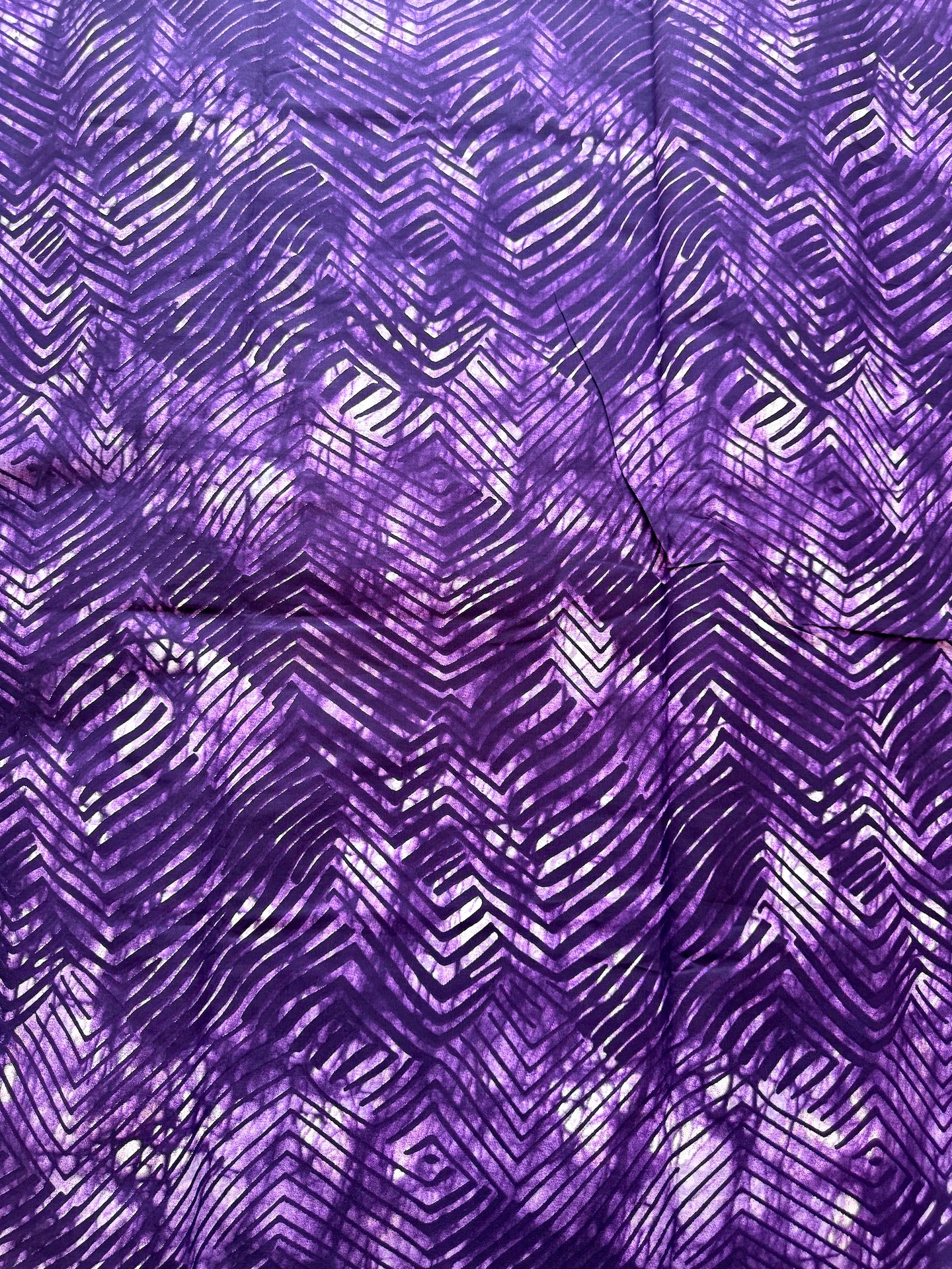 A close-up of a purple batik fabric with a hand-drawn chevron and diamond pattern in shades of purple and white.