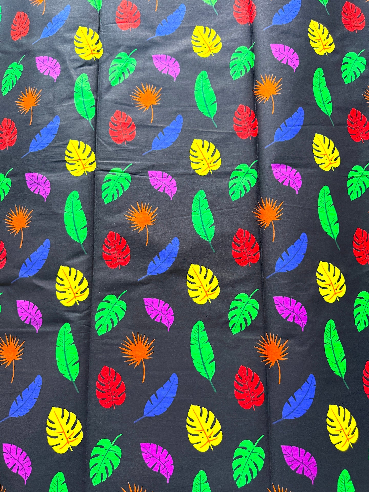A close-up of a dark charcoal gray fabric printed with a colorful tropical leaf pattern featuring green, yellow, red, blue, purple, and orange leaves.