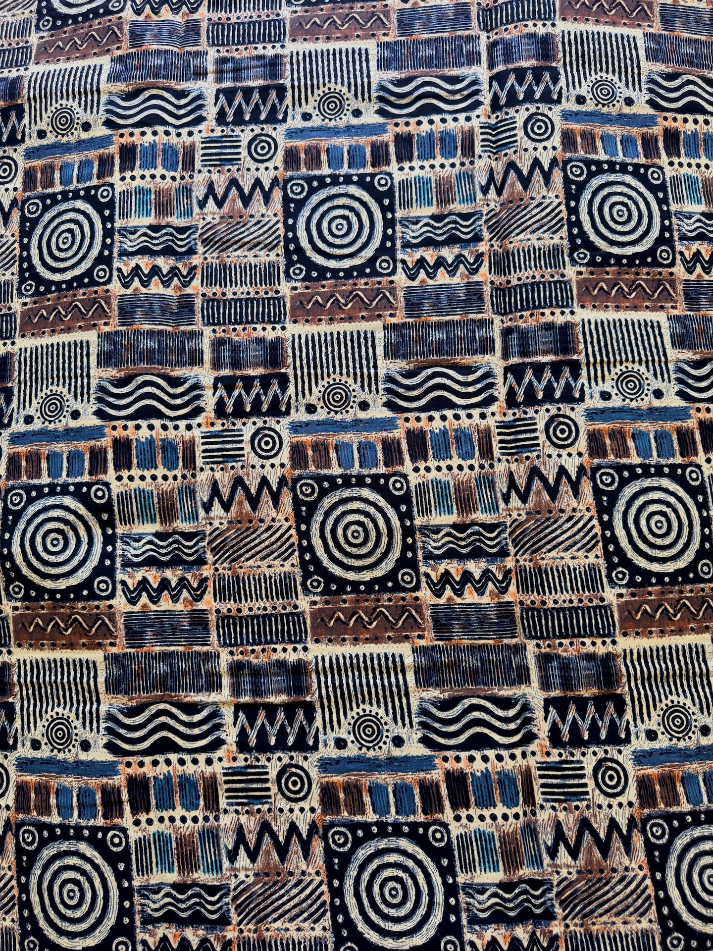A close-up of a textile or wallpaper pattern featuring a patchwork of geometric and tribal-inspired motifs in navy blue, brown, black, and off-white.