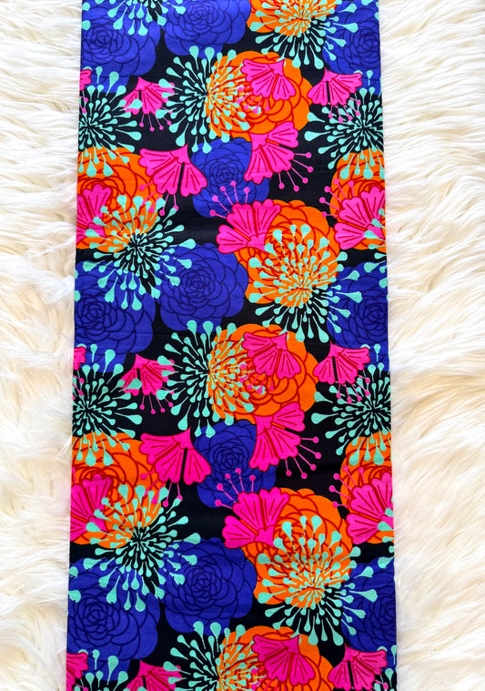A long rectangular fabric item, likely a scarf or narrow bandana, featuring a bold retro floral pattern in vibrant pink, orange, blue, and teal on a black background, laid flat on a white faux fur surface.