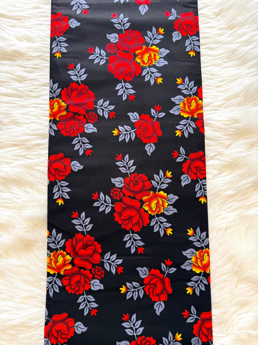 A long rectangular fabric runner with a bold floral print, laid flat on a white faux fur surface. The print features large red and yellow roses with gray leaves on a black background.