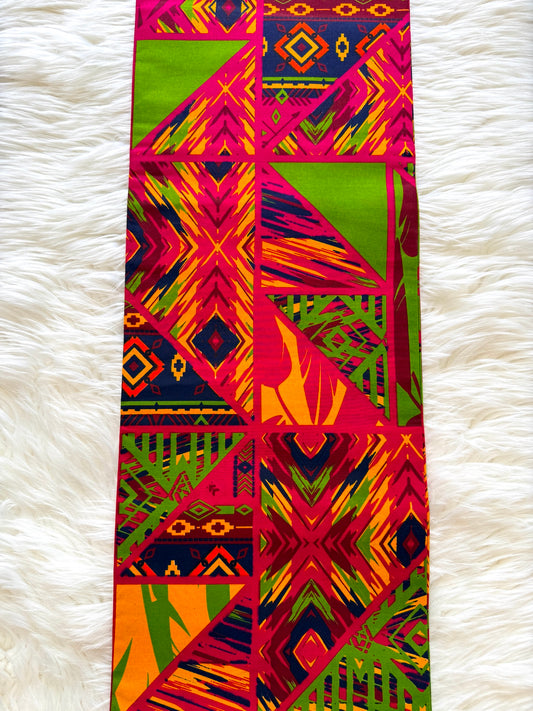 A long rectangular textile with a bold geometric and tribal-inspired print in bright pink, lime green, orange, yellow, and dark navy, laid flat on a white faux-fur surface.
