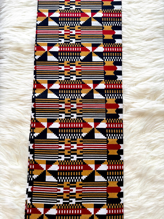 A long rectangular textile with a bold geometric pattern in black, red, white, and mustard yellow, laid flat on a white faux-fur surface.