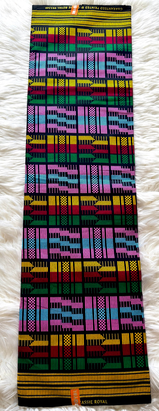 A folded multicolor African wax print fabric garment, likely a skirt or dress, laid flat on a white faux fur surface. The garment features a vibrant, repeating geometric pattern with stripes of pink, yellow, green, blue, red, and black.
