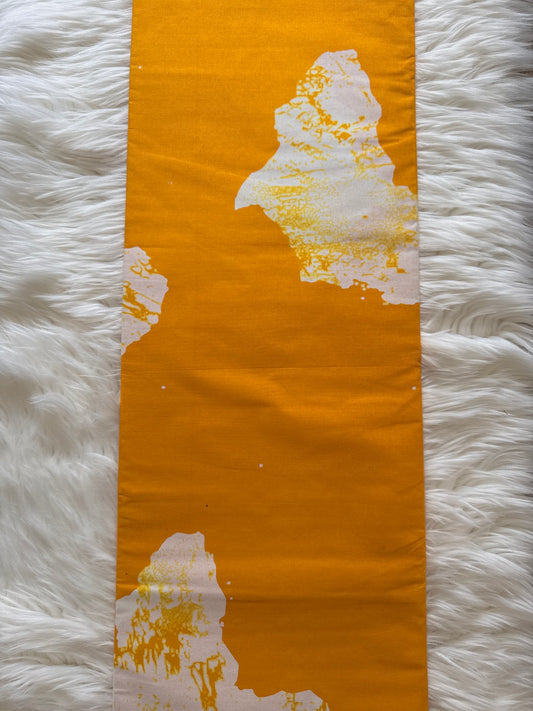 A bright orange fabric runner with white abstract painted patches, laid flat on a white faux fur surface.