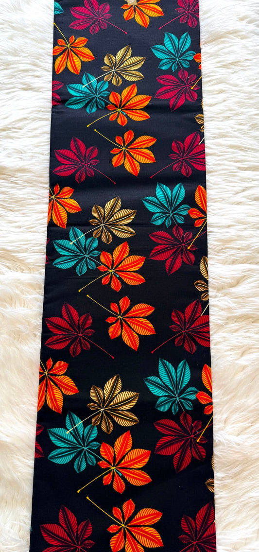 A long rectangular textile laid flat on a white faux-fur surface, featuring a bold multicolor floral pattern with orange, red, teal, and gold leaf-like motifs on a black background.