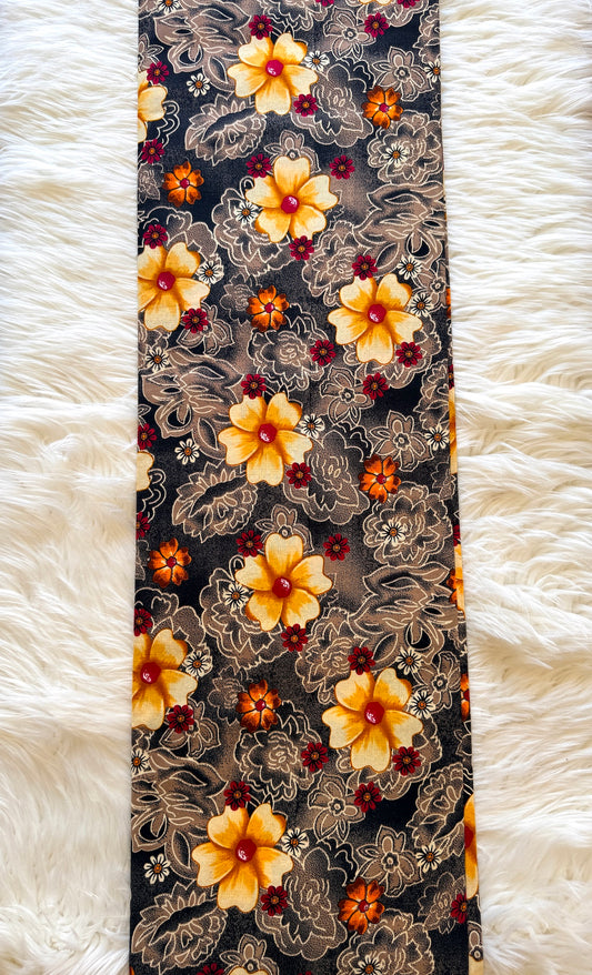 A long rectangular fabric item, likely a scarf or bandana, featuring a dense floral and ornamental print in yellow, orange, red, and beige on a black background, laid flat on a white faux fur surface.