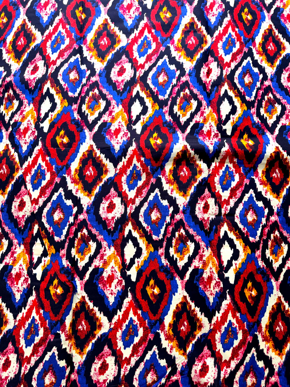 A close-up of a colorful geometric textile pattern with repeating diamond motifs in red, blue, pink, yellow, and white on a dark background.