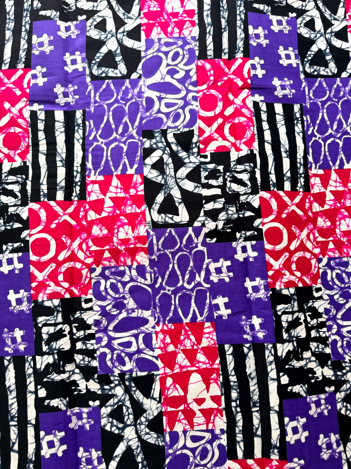 A close-up of a batik fabric featuring a patchwork design of purple, pink, black, and white patterns including stripes, crosses, and abstract shapes.