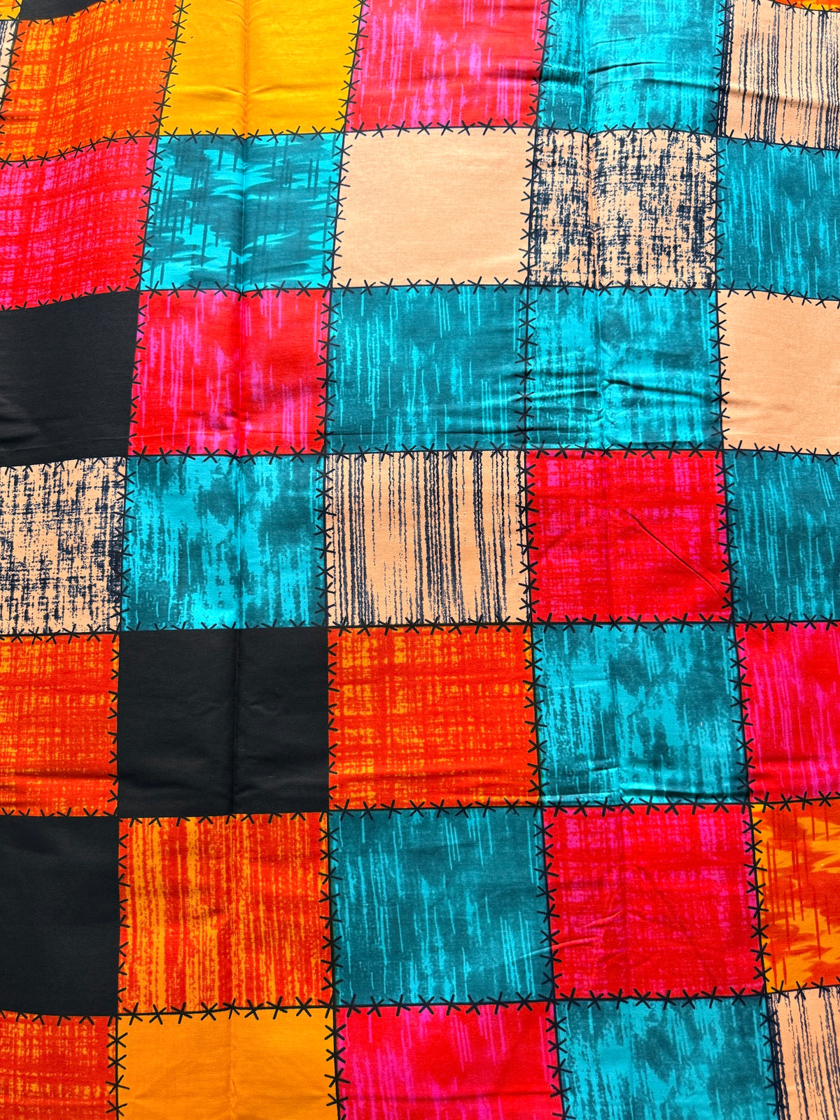 A close-up of a colorful patchwork fabric with stitched square panels in bright orange, turquoise, pink, black, beige, and yellow.