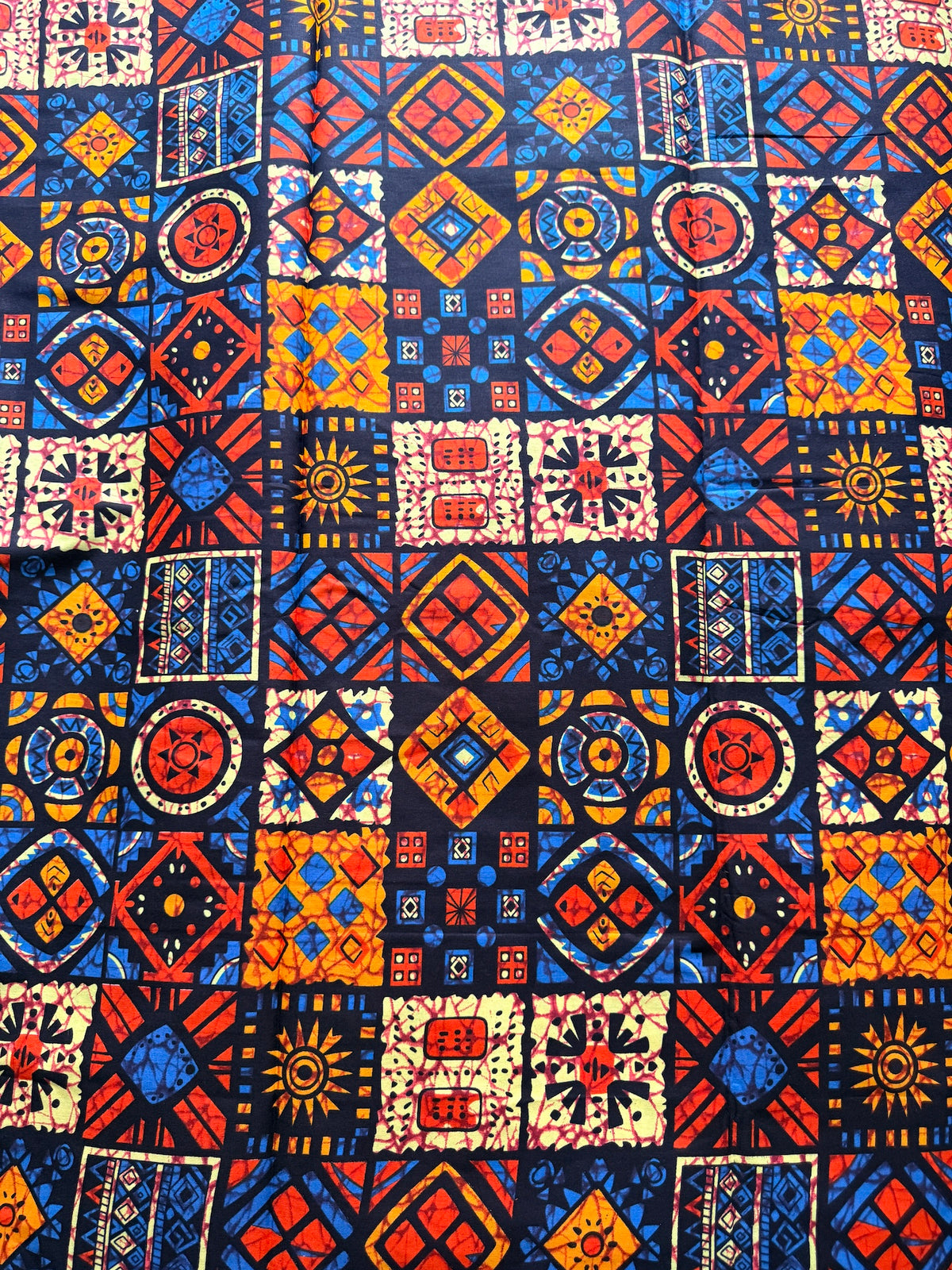 A close-up of a vibrant, multicolor African wax print fabric featuring a patchwork grid of geometric and tribal patterns in red, blue, yellow, and cream.