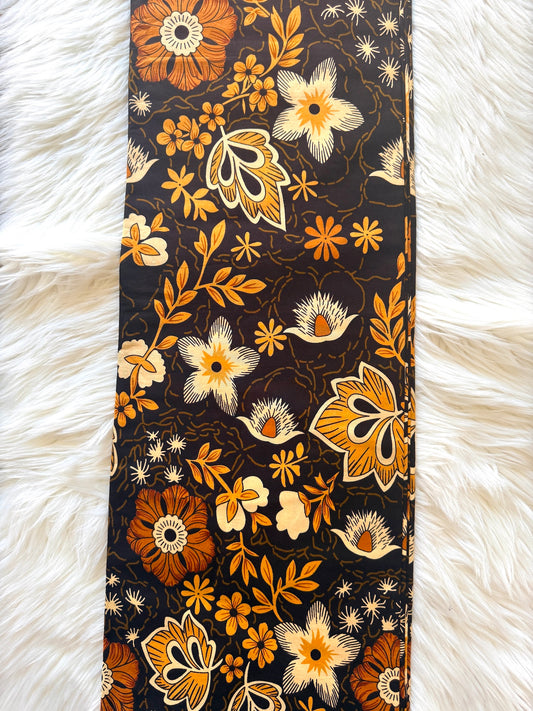A long rectangular scarf laid flat on a white faux-fur surface, featuring a dense floral and botanical print in orange, mustard yellow, cream, and brown on a very dark charcoal background.