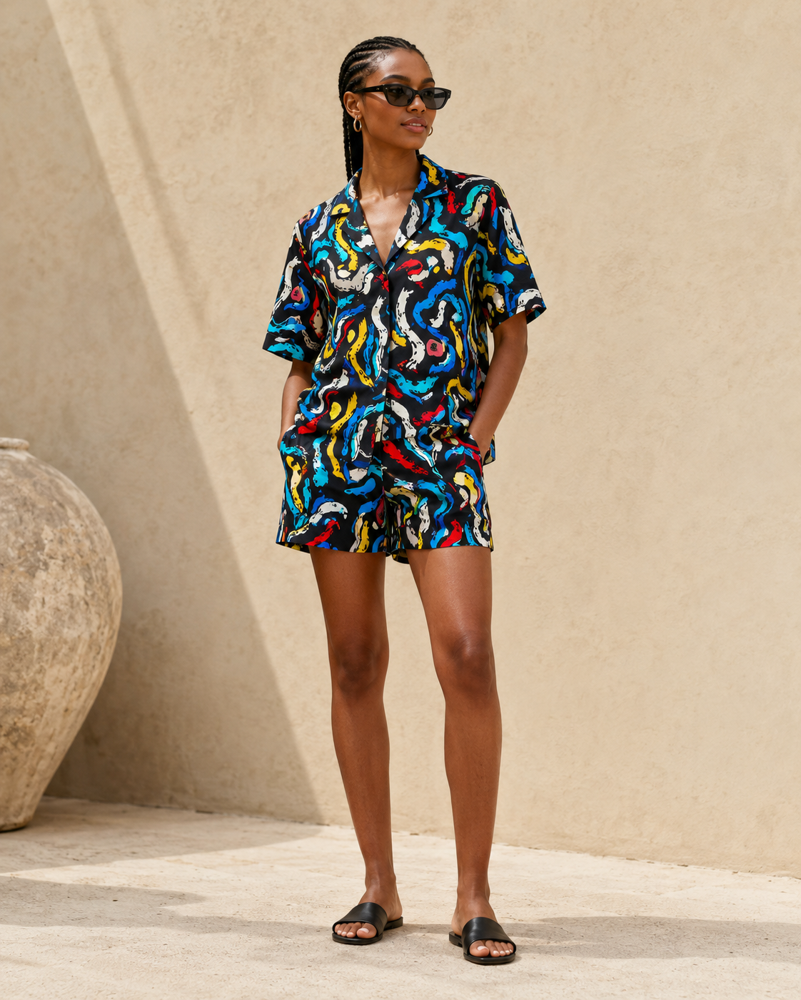 A woman modeling a matching short-sleeve button-up shirt and shorts set with a vibrant, multicolored abstract wavy print, standing against a beige stucco wall.
