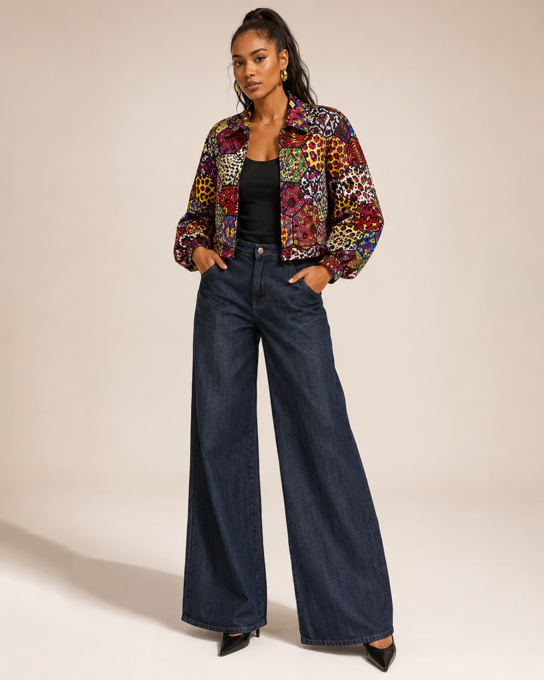 A woman modeling a colorful patterned cropped jacket over a black top and dark blue wide-leg jeans, standing against a plain beige studio background.