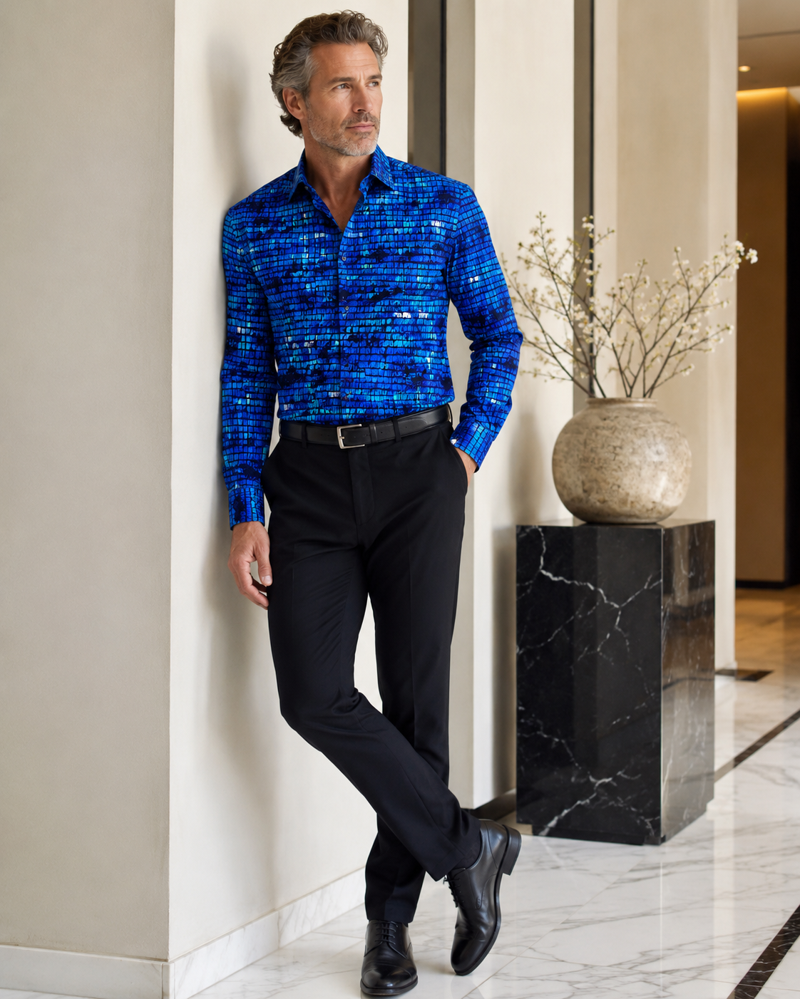Man in a blue patterned shirt and black pants leaning against a wall in an elegant interior setting.