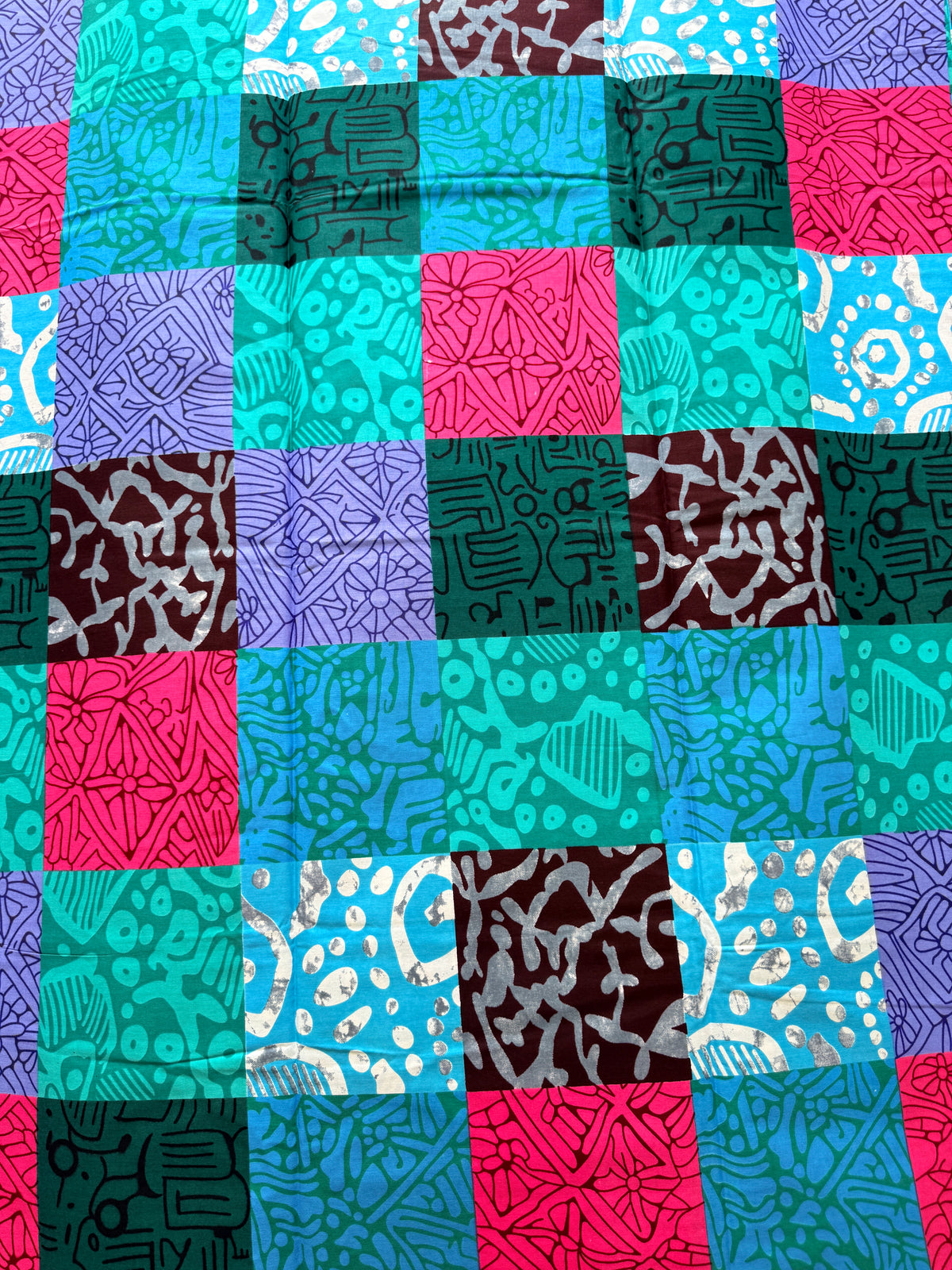 A close-up of a patchwork quilt or fabric panel made of square blocks in bright turquoise, teal, pink, purple, dark green, and brown, each with different abstract line and dot patterns.
