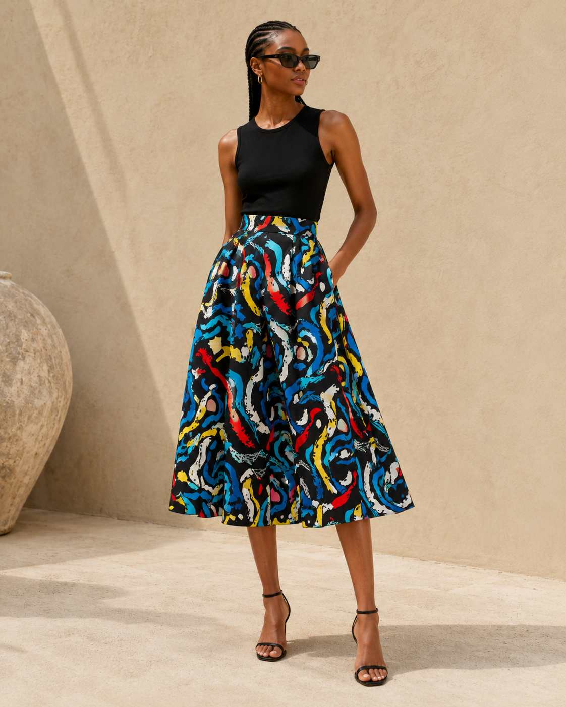 A woman modeling a sleeveless black top and a high-waisted midi skirt with a bold, multicolored abstract swirl pattern in blue, red, yellow, and white, set against a beige textured wall.