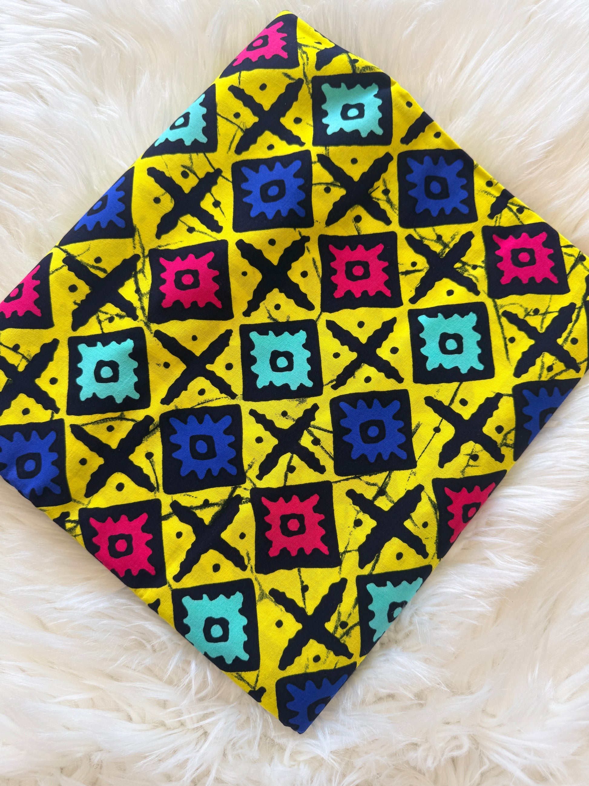 A square fabric bandana or scarf with a vibrant yellow background and a colorful geometric pattern, laid flat on a white faux fur surface.