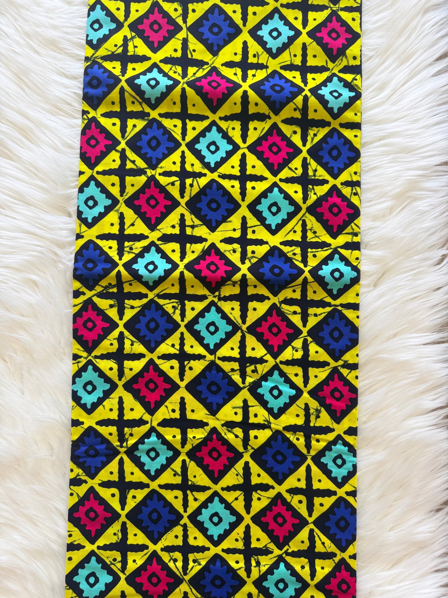 A rectangular textile laid flat on a white faux-fur surface, featuring a bold geometric diamond pattern in yellow, black, blue, pink, and turquoise.
