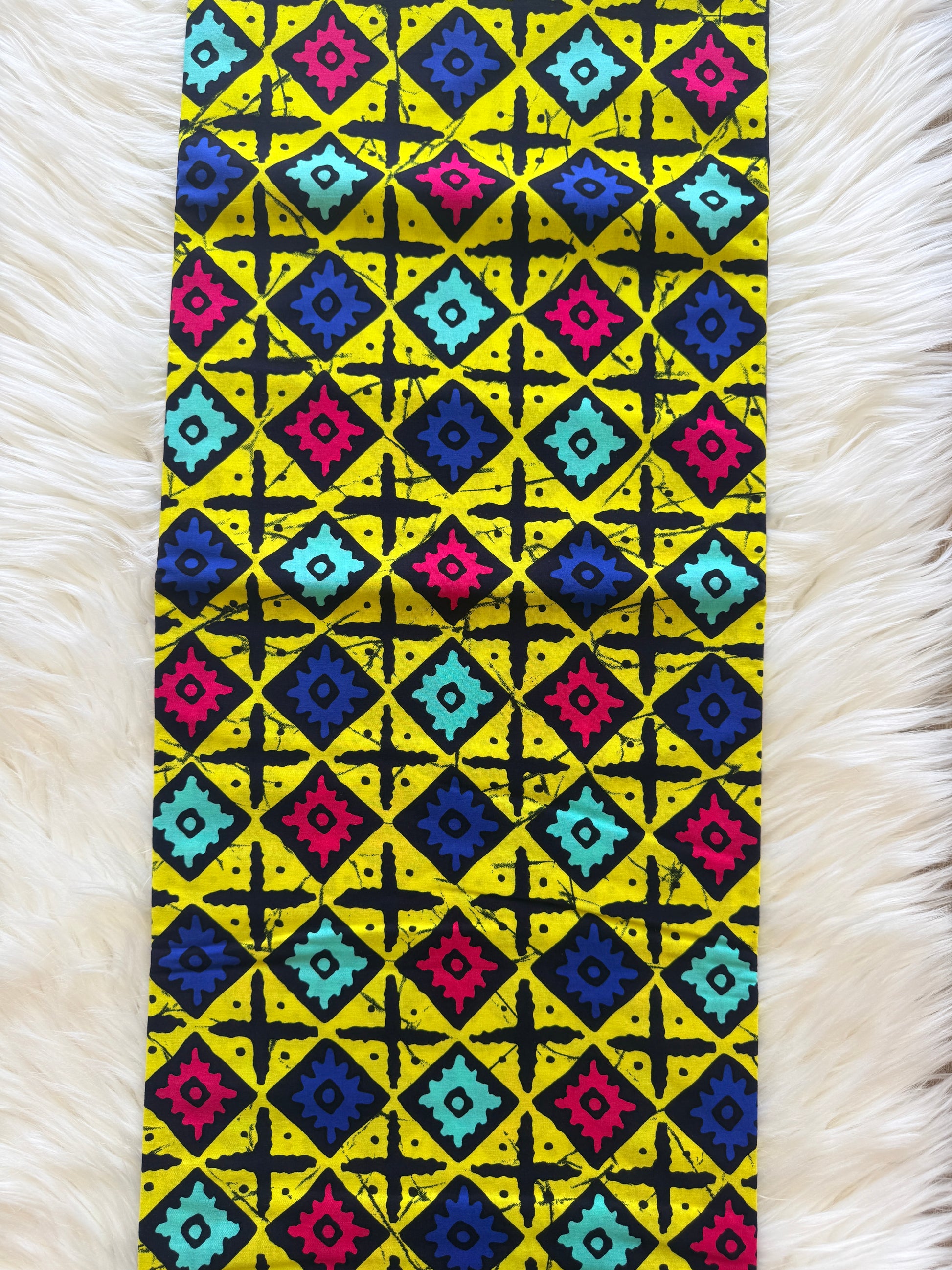 A rectangular textile laid flat on a white faux-fur surface, featuring a bold geometric diamond pattern in yellow, black, blue, pink, and turquoise.