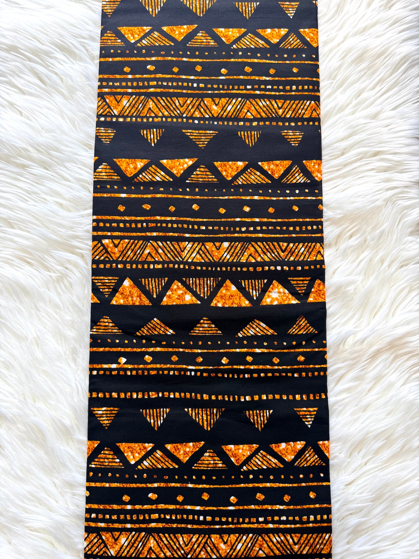 A long rectangular textile with an African wax print pattern in black and orange, laid flat on a white faux fur background.