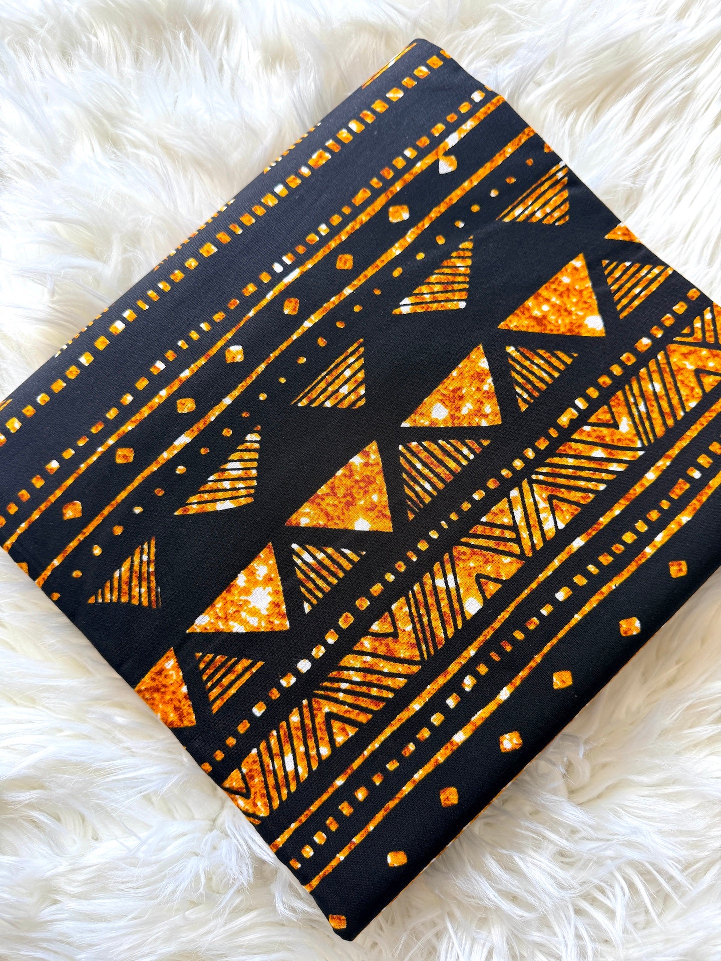 A square fabric item, likely a scarf or small bag, featuring a black base with a vibrant orange glitter geometric pattern of triangles, stripes, and dots, laid flat on a white faux fur surface.