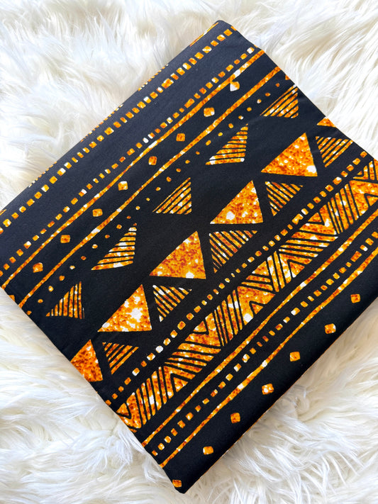 A square fabric item, likely a scarf or small bag, featuring a black base with a vibrant orange glitter geometric pattern of triangles, stripes, and dots, laid flat on a white faux fur surface.