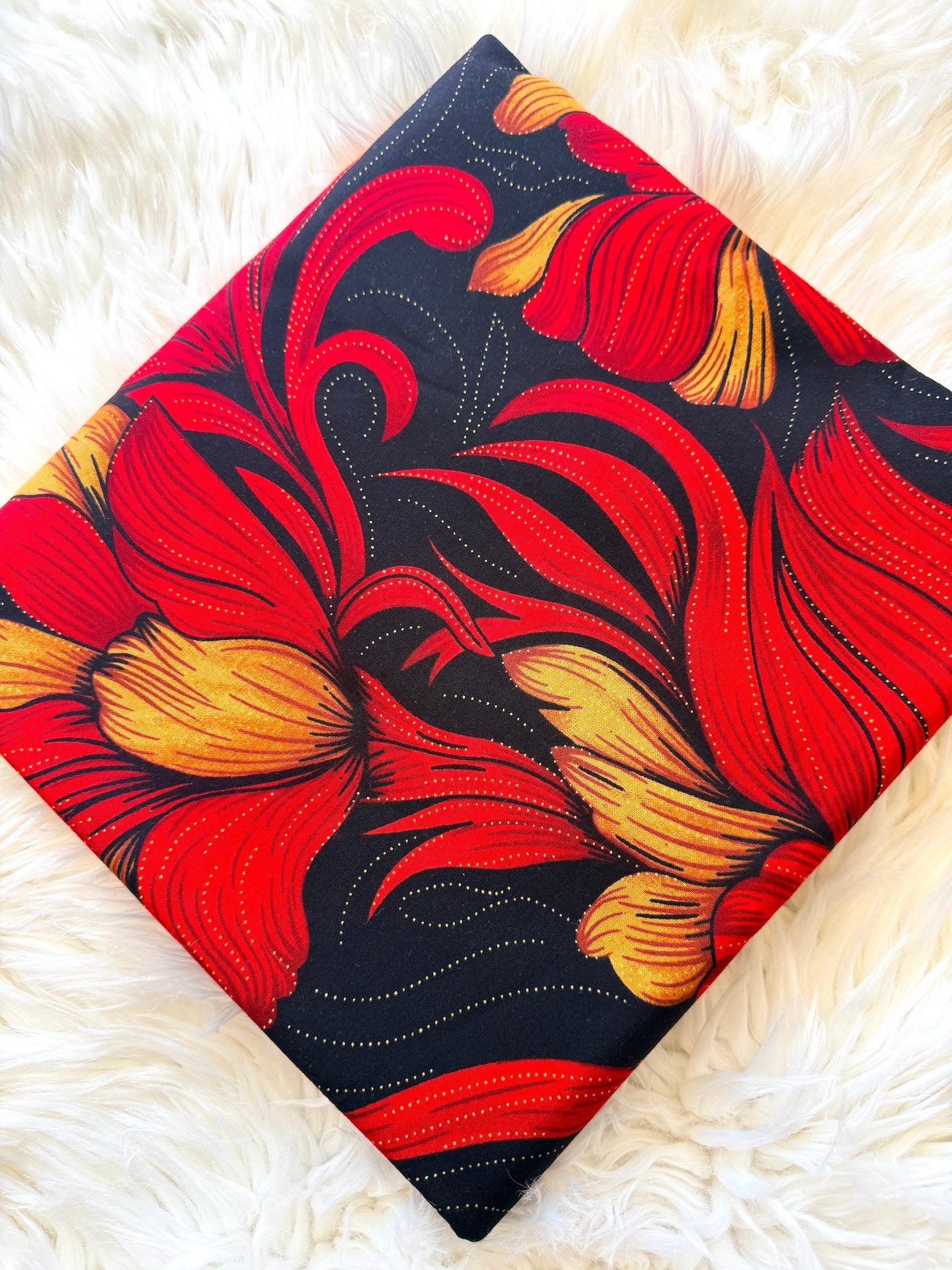 A square fabric item, likely a pillow cover or small blanket, featuring a bold floral pattern in vibrant red and yellow on a black background, laid flat on a white faux fur surface.