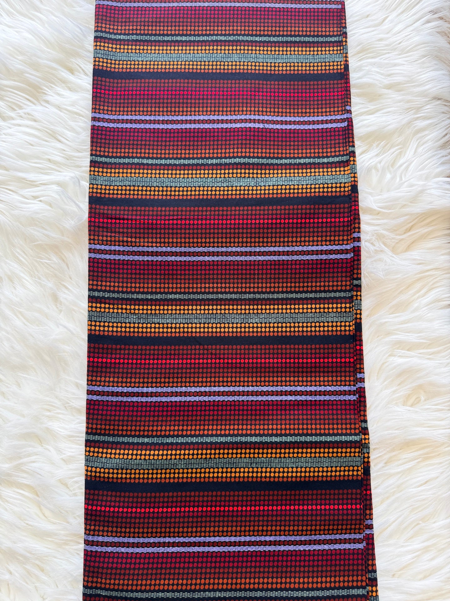 A striped woven textile, likely a scarf or narrow fabric panel, featuring horizontal bands of red, black, orange, yellow, and light blue on a white faux fur background.