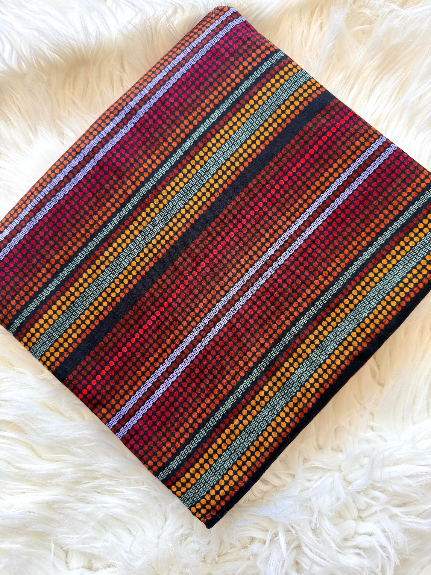 A folded woven textile with a multicolor striped pattern of red, orange, yellow, black, and white, displayed on a white faux fur surface.