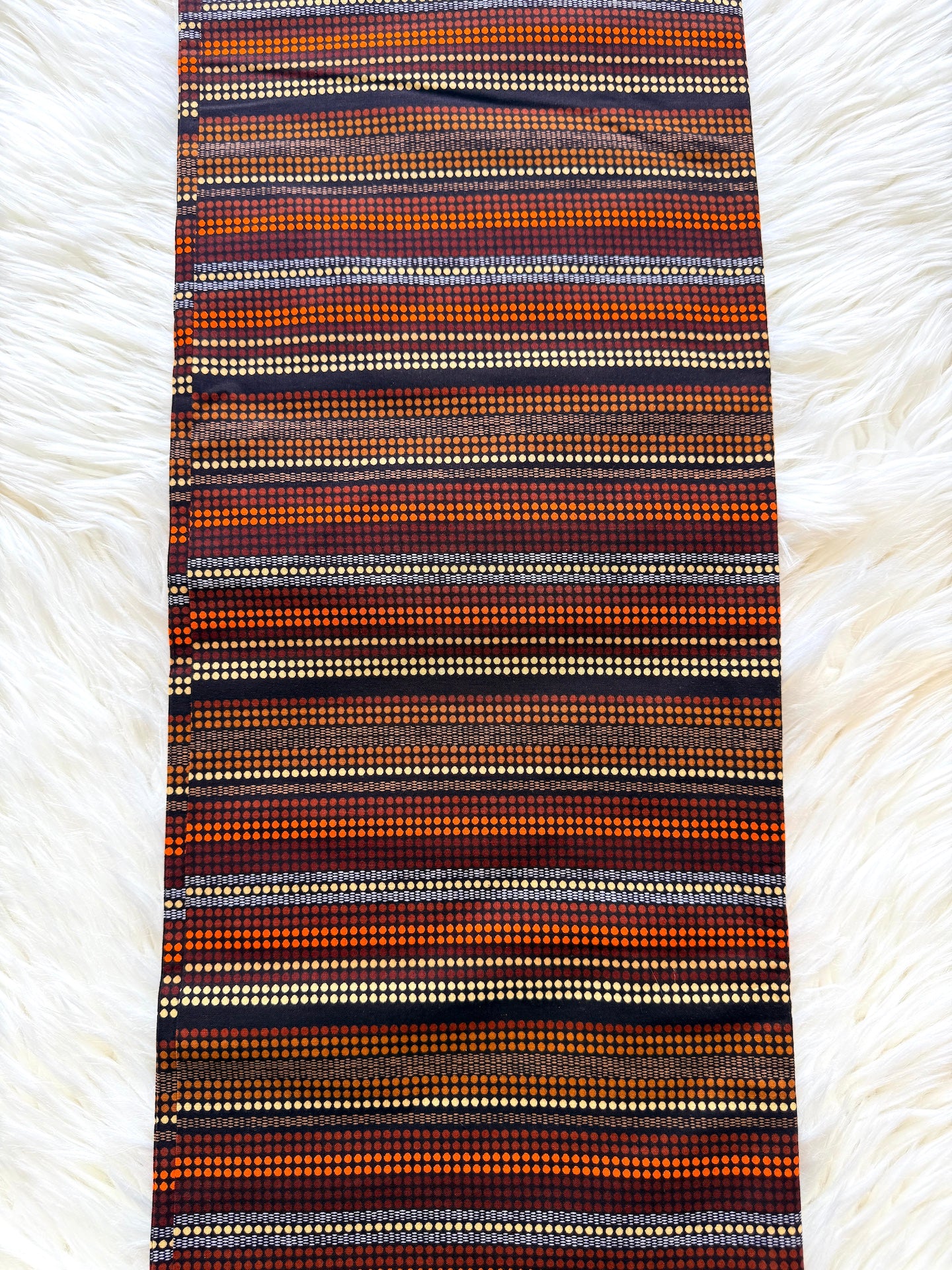 A long rectangular woven textile with horizontal striped patterns in orange, brown, black, beige, and white, laid flat on a white faux fur surface.