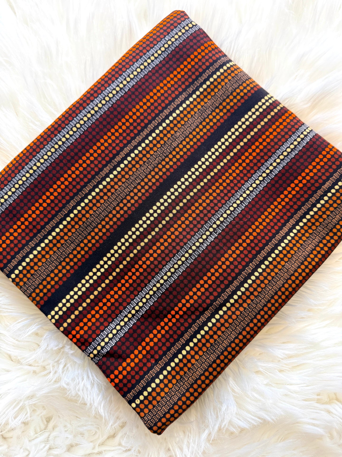 A square throw pillow with a detailed geometric striped pattern in orange, brown, red, black, and cream, displayed on a white faux fur surface.