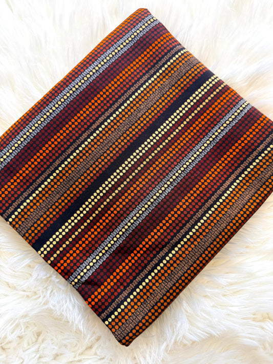 A square throw pillow with a detailed geometric striped pattern in orange, brown, red, black, and cream, displayed on a white faux fur surface.