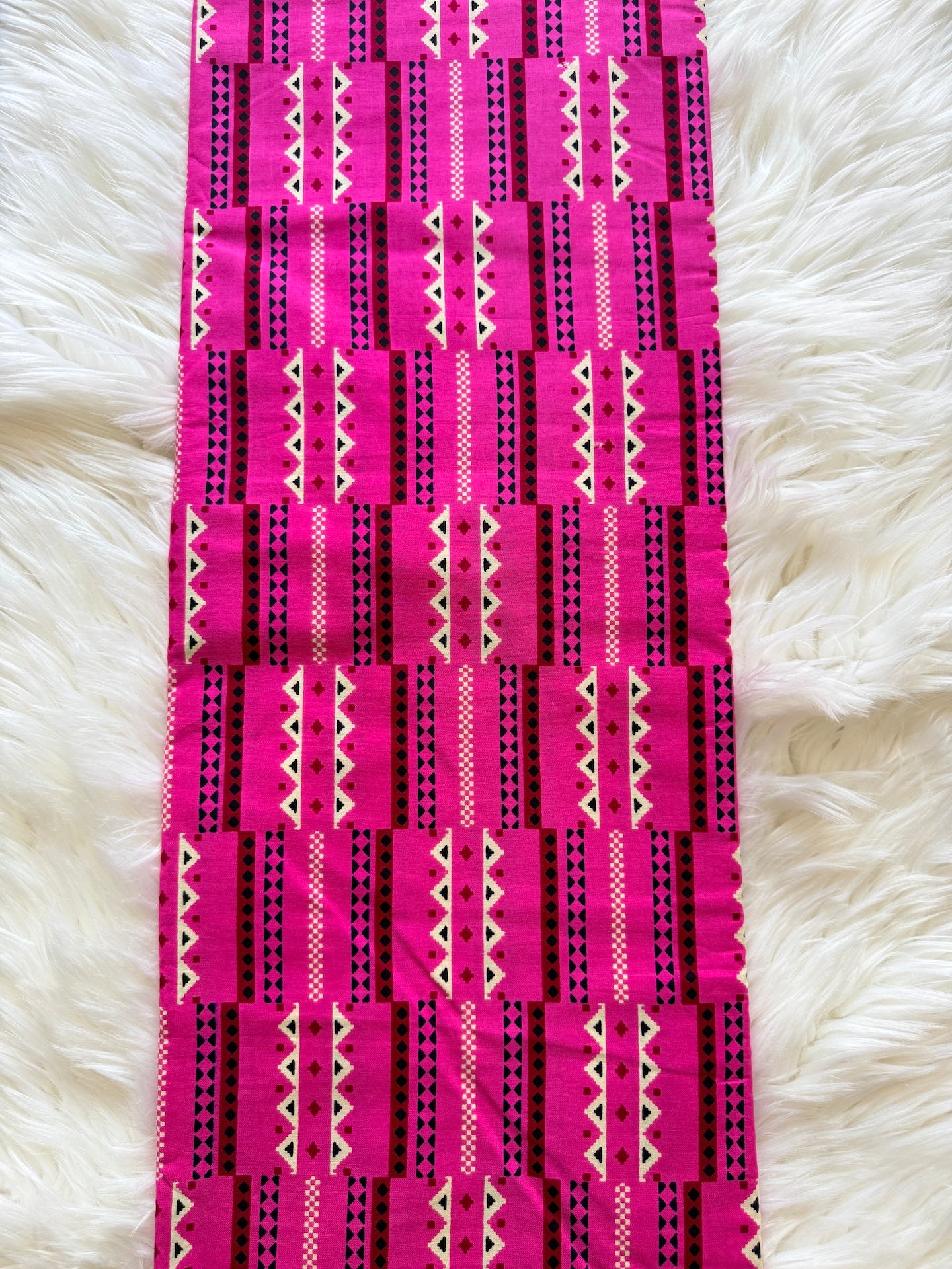 A bright pink and black patterned fabric runner or table runner laid flat on a white faux fur surface. The fabric features geometric tribal-style patterns with vertical stripes, including pink, black, and cream zig-zag and dotted designs.