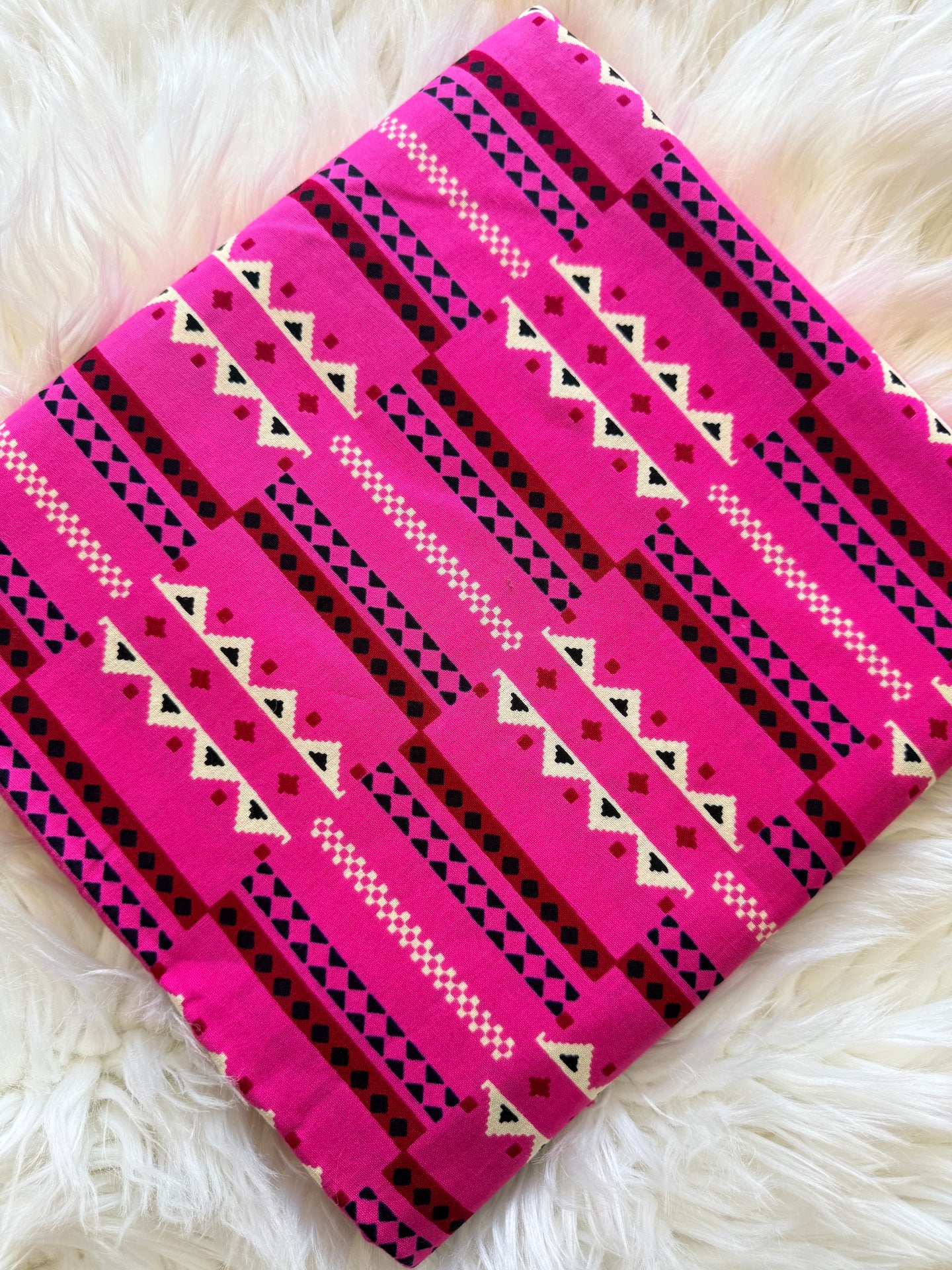 A folded textile item, likely a blanket or throw, featuring a bright pink and black Southwestern geometric pattern with white accents, laid flat on a white faux fur surface.