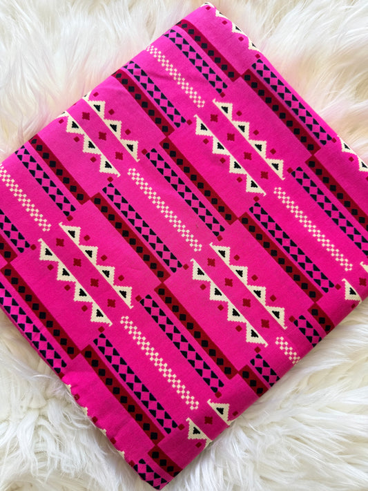 A folded textile item, likely a blanket or throw, featuring a bright pink and black Southwestern geometric pattern with white accents, laid flat on a white faux fur surface.