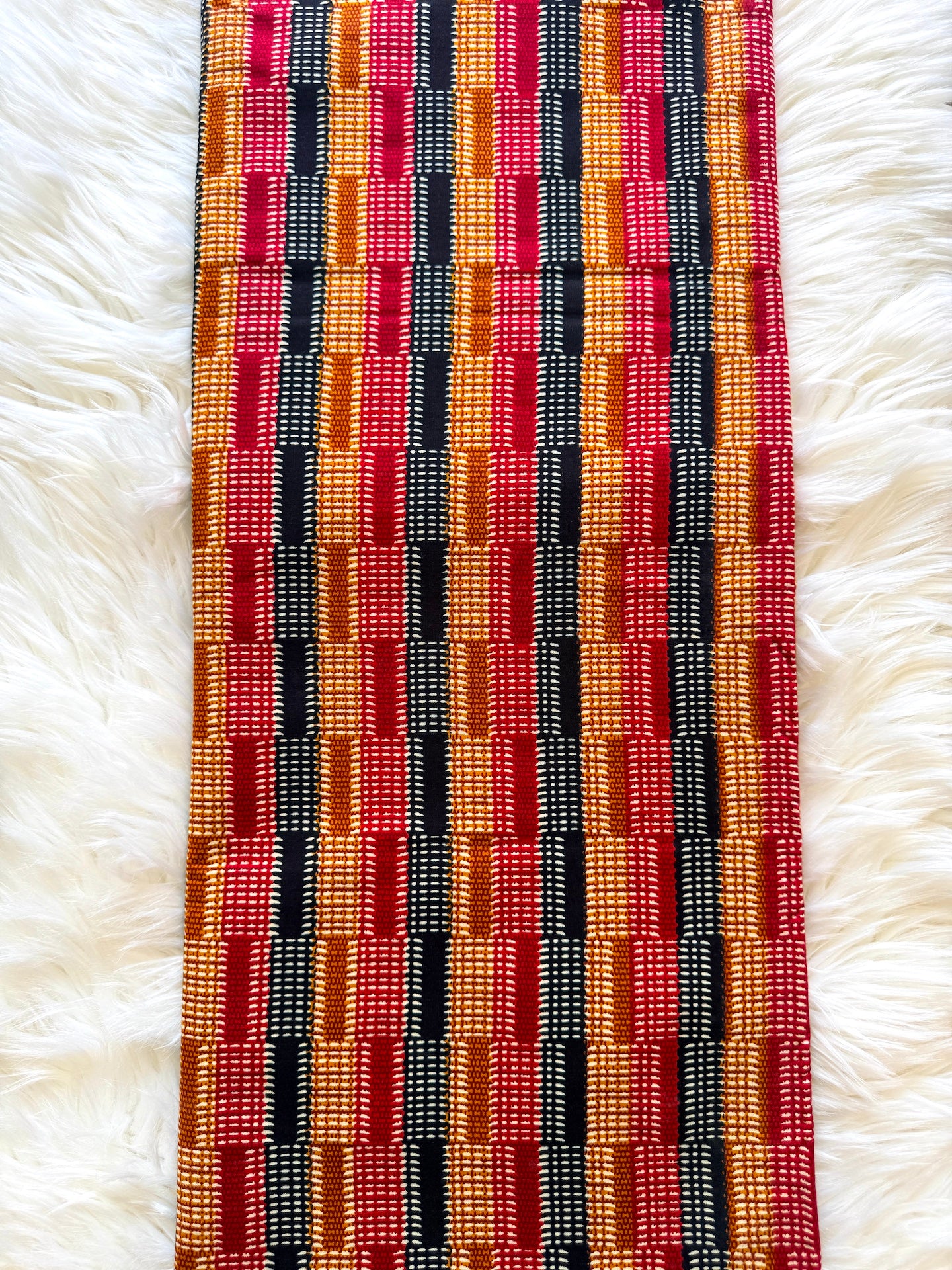A long rectangular woven textile with a bold geometric pattern in red, orange, black, and white, laid flat on a white faux fur surface.