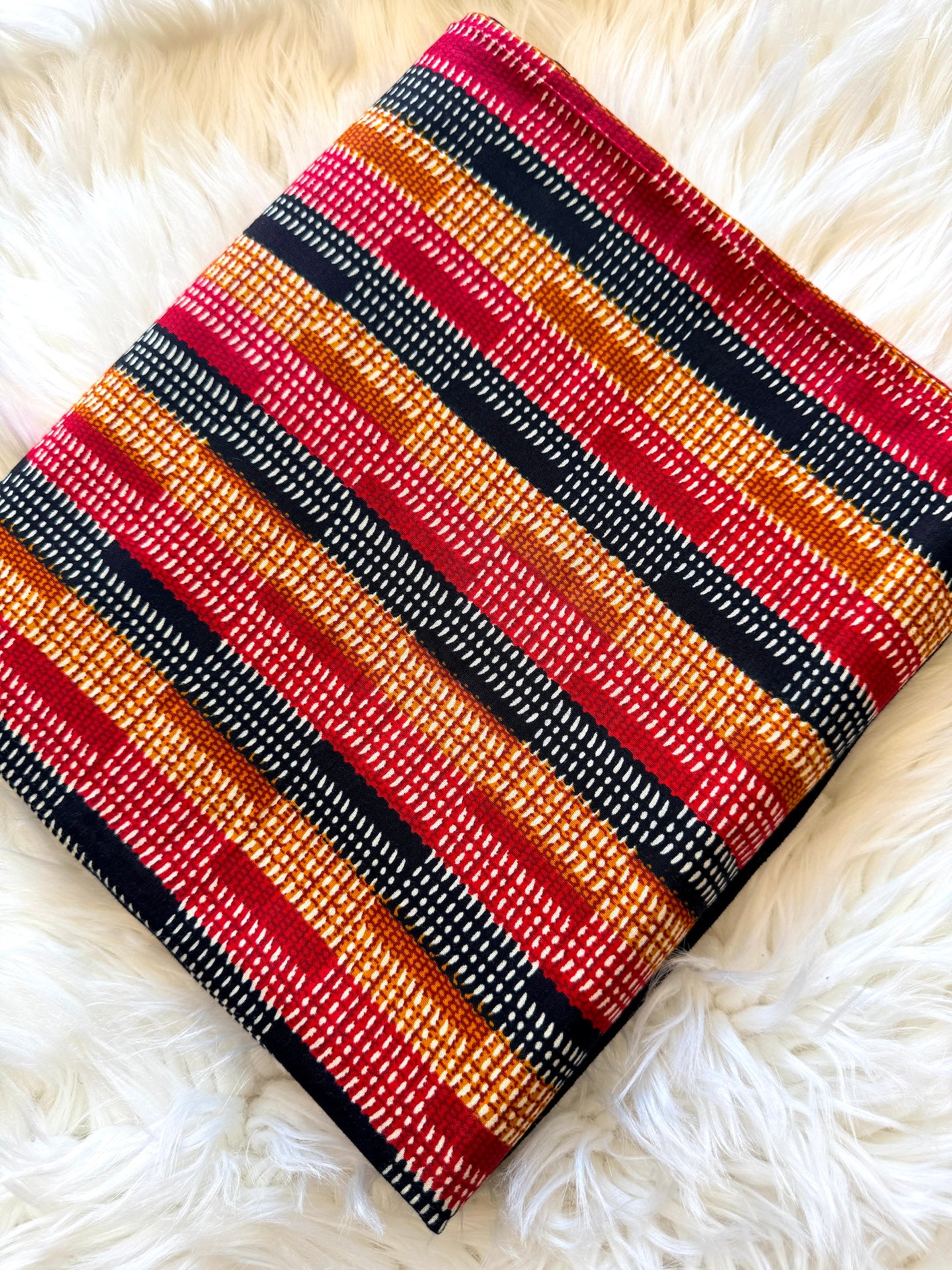 A square woven textile or pouch with a bold striped pattern in red, orange, black, and white, laid flat on a white faux fur surface.