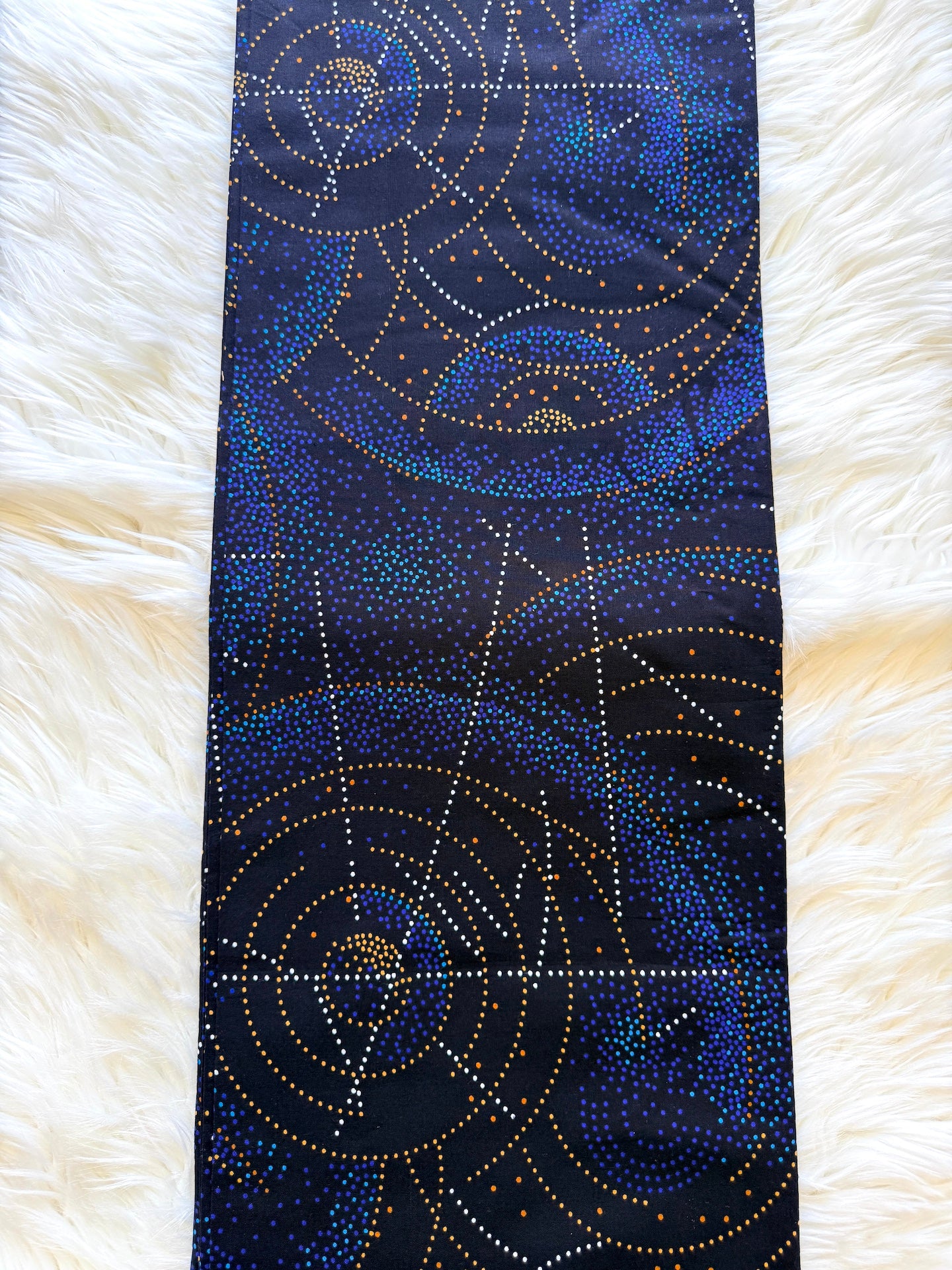A long, narrow scarf laid flat on a white faux fur surface. The scarf has a dark navy blue background with a repeating pattern of gold, blue, and white dotted circular and geometric designs.