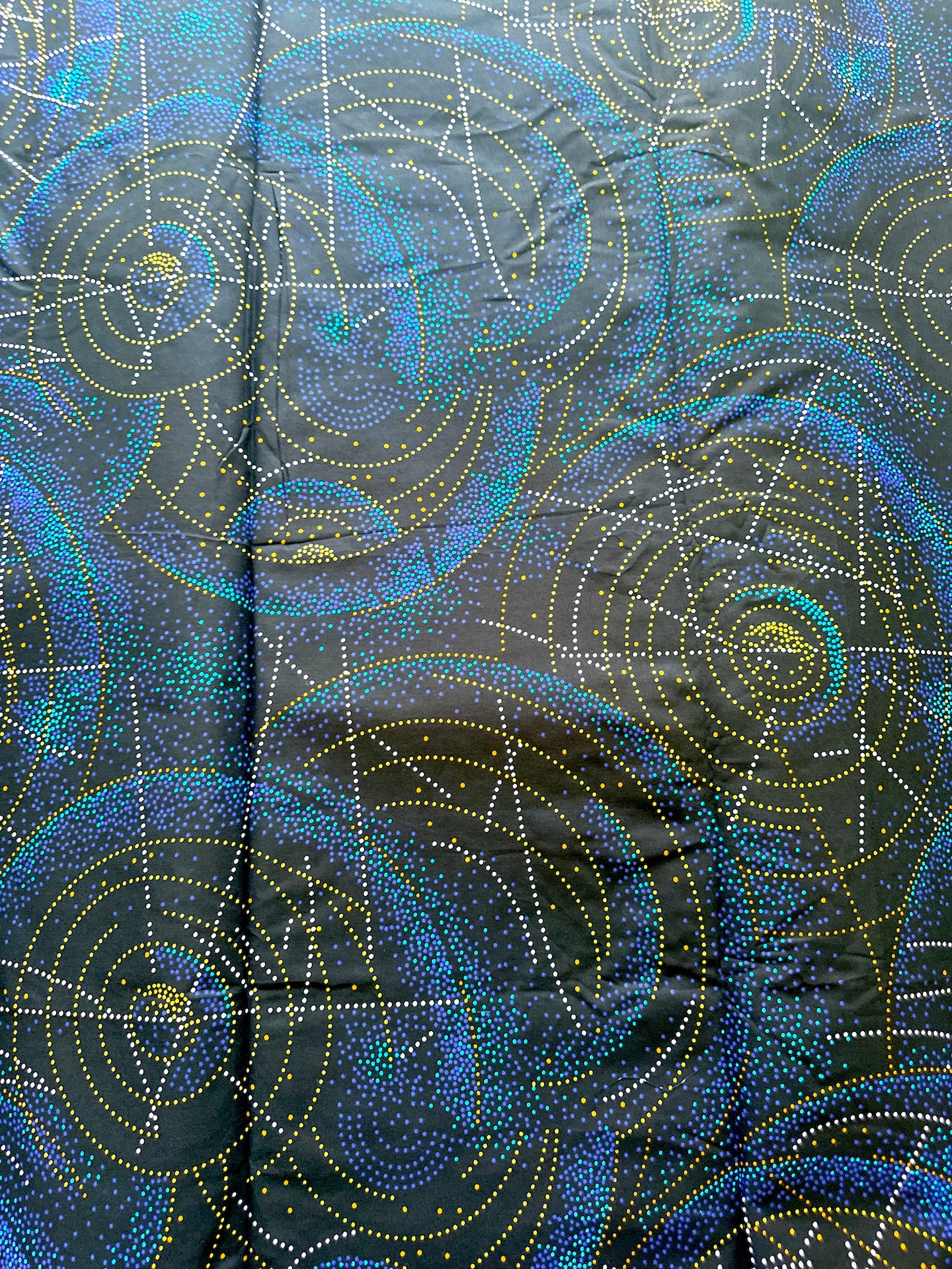 A close-up of a black fabric with a repeating cosmic pattern of blue, teal, and yellow dotted spiral galaxies and star fields.