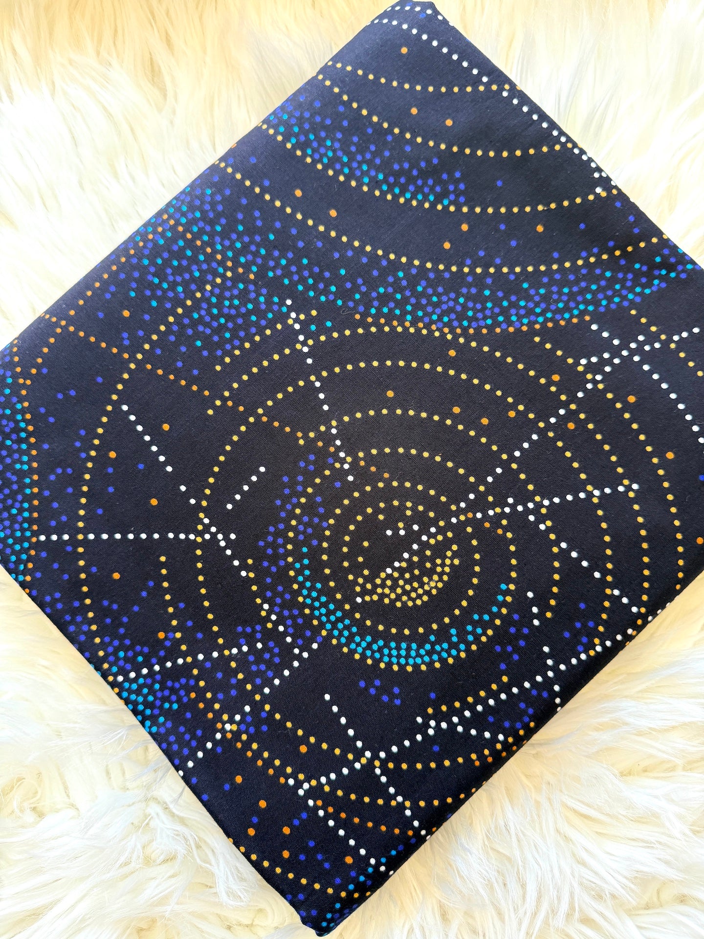 A square fabric item, likely a scarf or small bag, featuring a dark navy blue background with a circular dotted pattern in gold, blue, and white, displayed on a fluffy white faux fur surface.