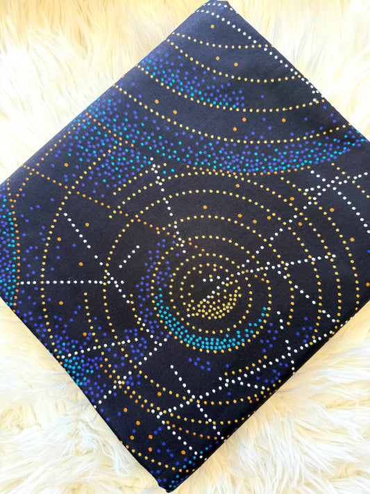 A square fabric item, likely a scarf or small bag, featuring a dark navy blue background with a circular dotted pattern in gold, blue, and white, displayed on a fluffy white faux fur surface.