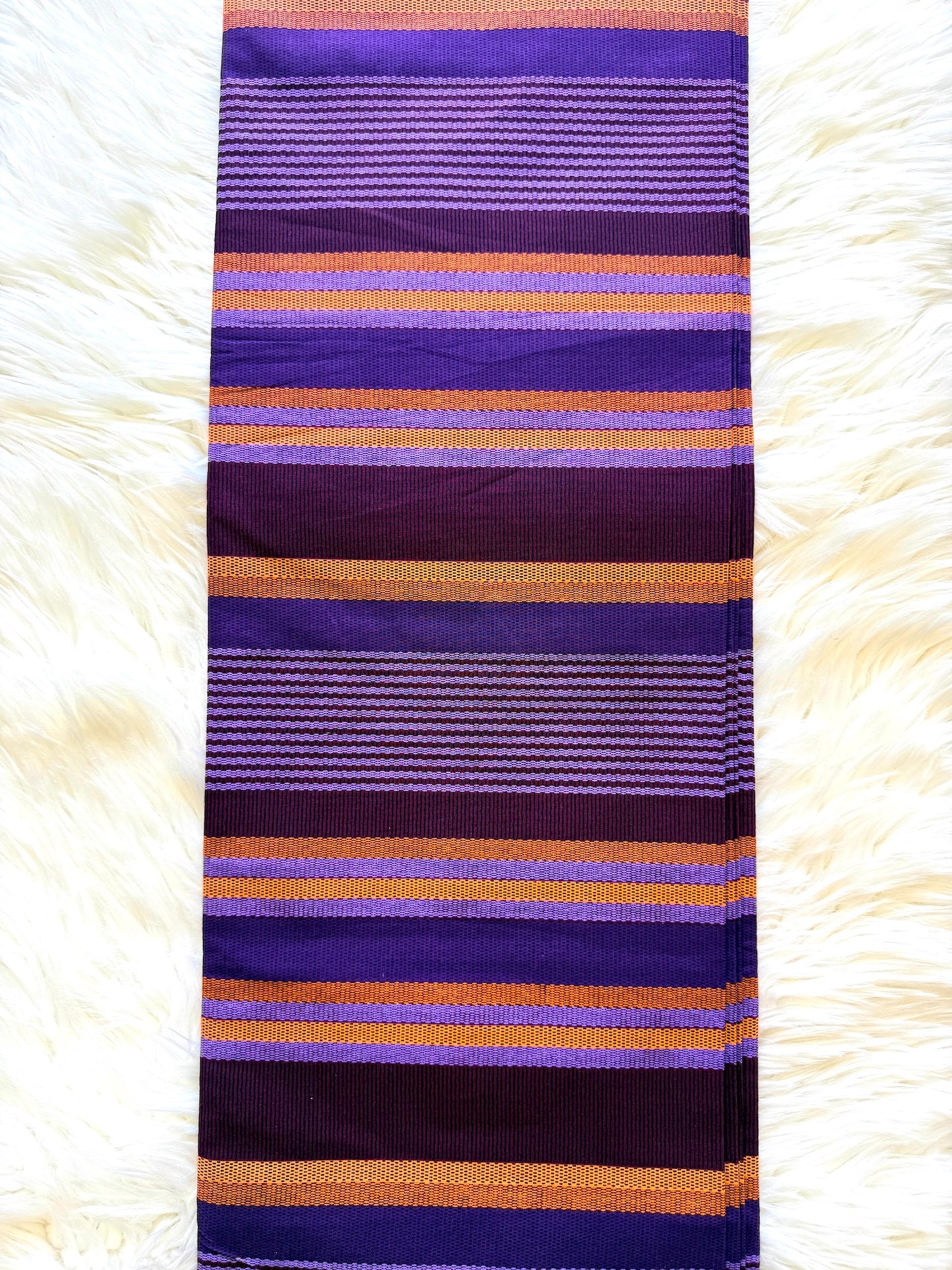 A striped fabric scarf or sash with horizontal bands of purple, violet, dark plum, and orange, laid flat on a white faux fur surface.
