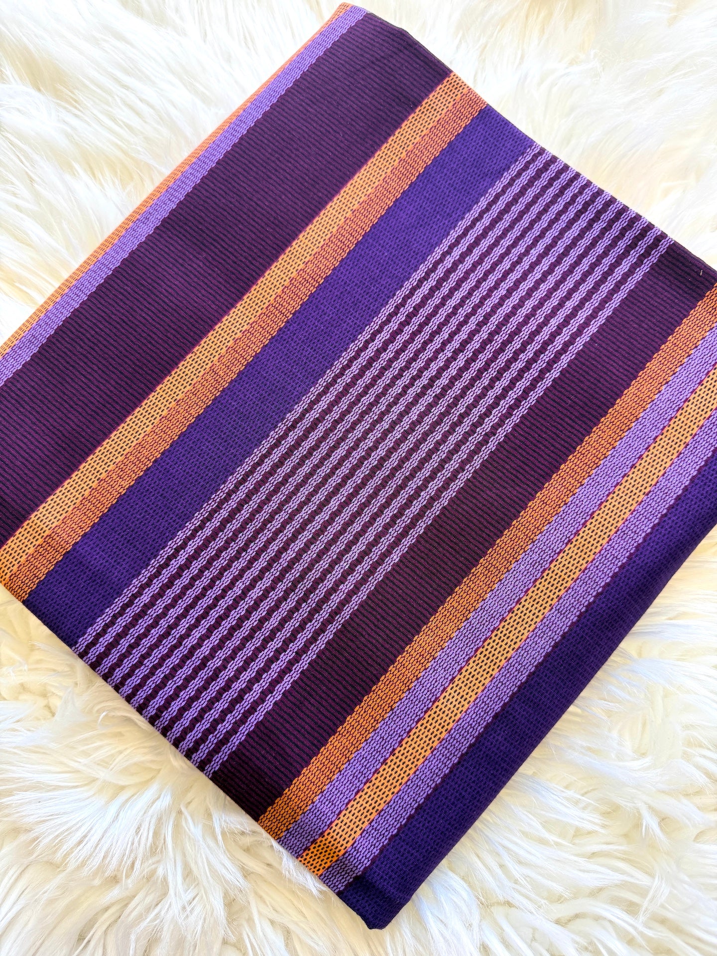 A square woven textile with horizontal purple, lavender, orange, and dark purple stripes, laid flat on a white faux fur surface.