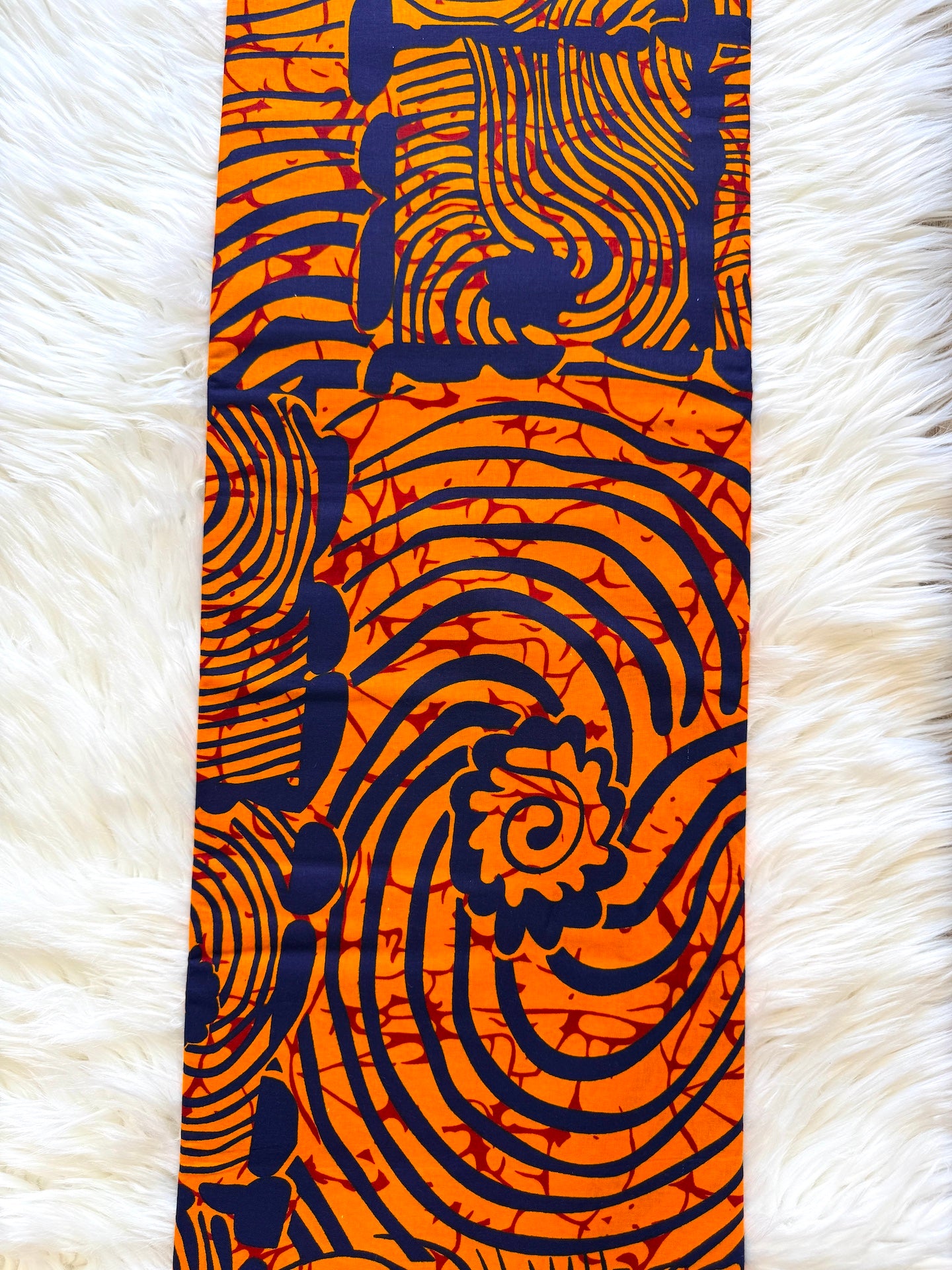 A vertical fabric panel featuring a bold orange and navy blue abstract pattern with swirls, stripes, and floral motifs, laid flat on a white faux fur background.