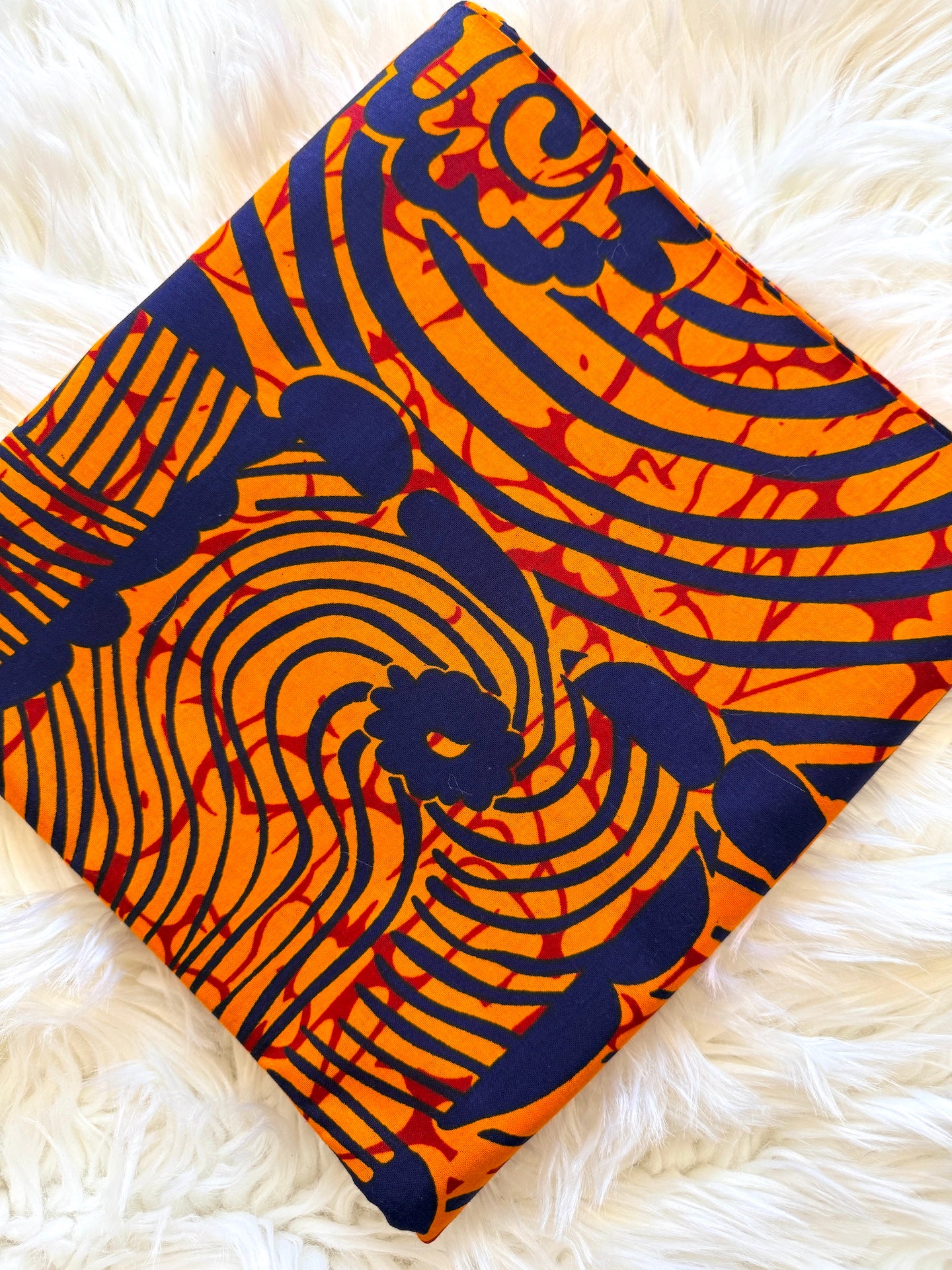 A square piece of vibrant orange and navy blue patterned fabric with red accents, laid flat on a white faux fur surface.