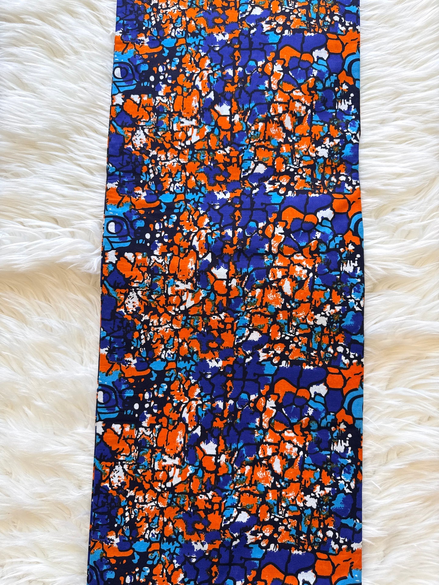 A rectangular fabric item, likely a scarf or bandana, featuring an abstract African wax print pattern in vibrant blue, orange, and white, laid flat on a white faux fur surface.