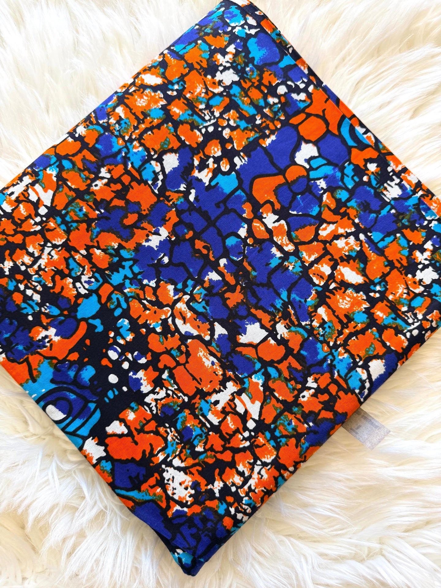 A square fabric item, likely a pillow cover or small blanket, featuring a vibrant abstract African wax print pattern in orange, blue, black, and white, laid flat on a white faux fur background.